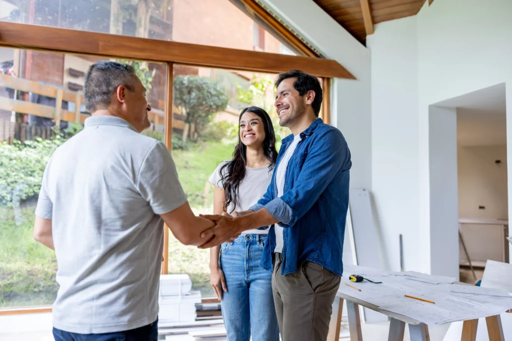 Two men shake hands while a woman observes in a well-lit room with large windows, surrounded by construction materials.