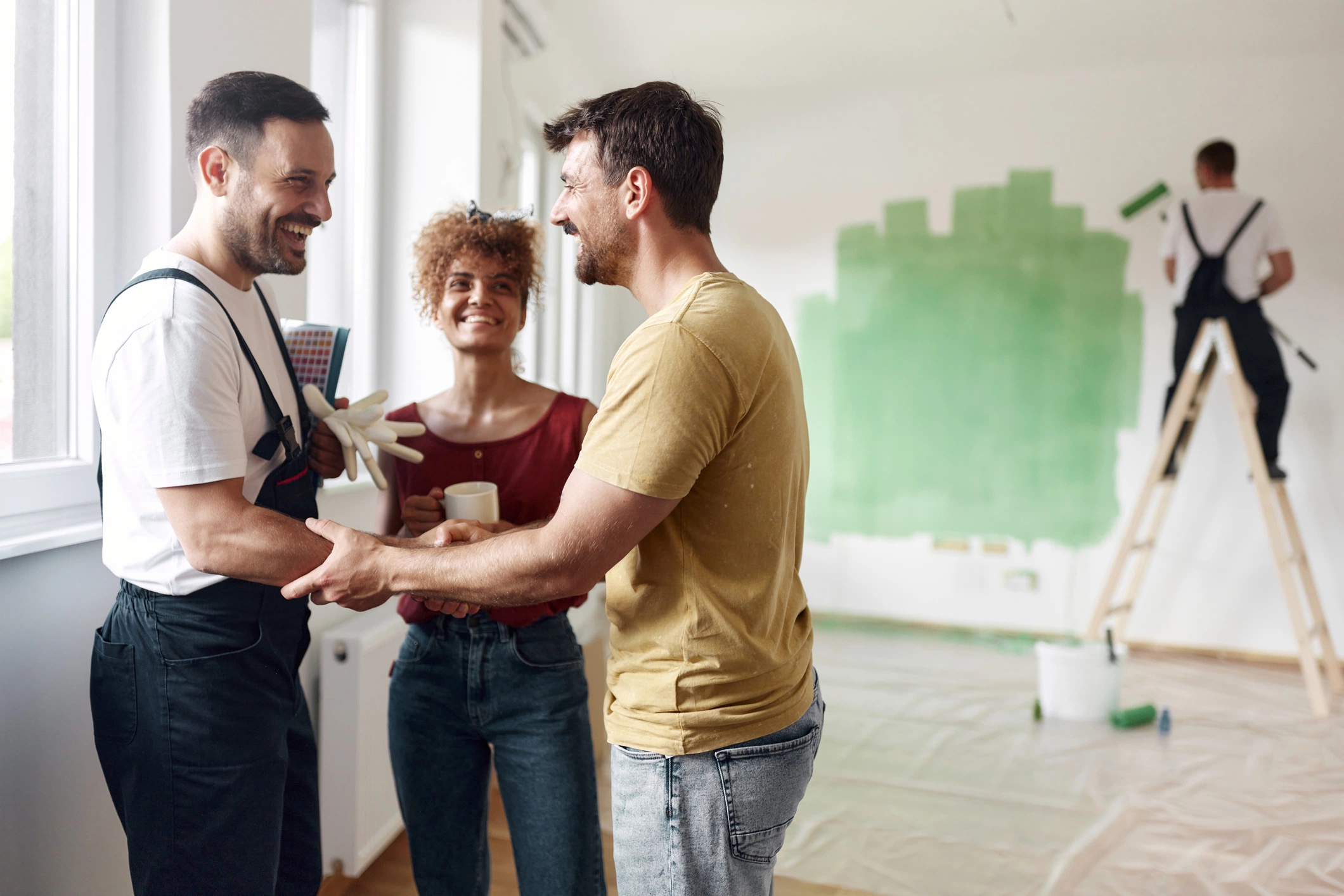 A couple shakes hands with a painter in a room being renovated, while another person paints a wall green in the background.
